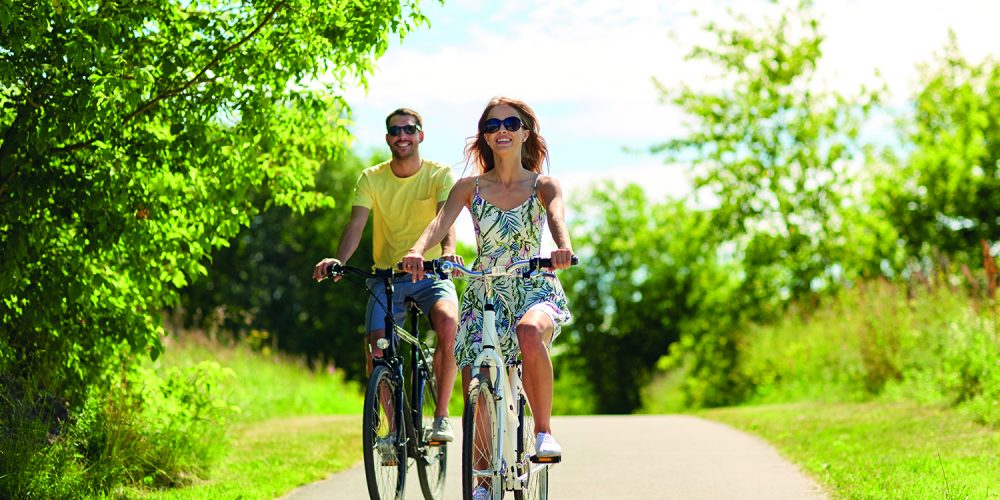 people, leisure and lifestyle concept - happy young couple riding bicycles along road in summer